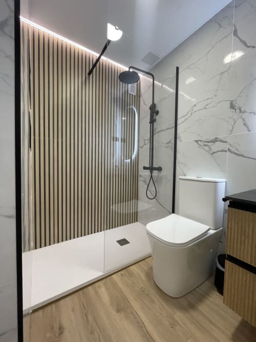 Baño moderno en Bahia Mar Canarias con ducha walk-in, alcachofa de lluvia, inodoro blanco, paredes de mármol y suelo de madera