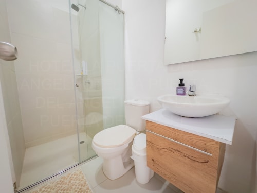 Baño moderno con lavabo tipo recipiente, vanidad de madera, azulejos blancos y ducha con mampara de vidrio en el hotel La Puerta del Angel
