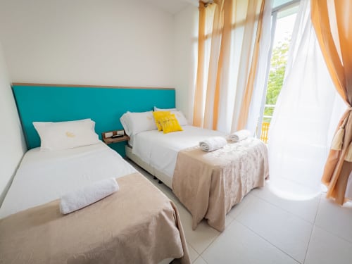 Luminosa habitación triple en hotel La Puerta del Angel en Ricaurte con cabecero turquesa, dos camas, sábanas blancas y jardín