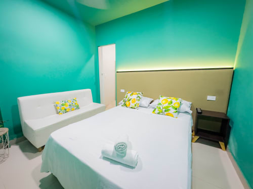 Dormitorio moderno con paredes turquesas, cama blanca, almohadas amarillas estampadas y zona de estar en el hotel La Puerta del Ángel