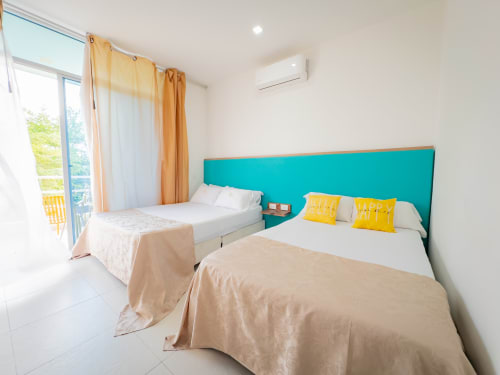 Luminosa habitación con dos camas dobles, cabecero turquesa, almohadas amarillas y balcón con vistas al río en La Puerta del Angel