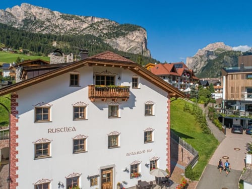 Rustclea Apartments hotel with white facade and wooden balcony in Selva di Valgardena, surrounded by Dolomite mountains