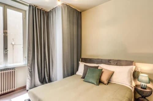 Camera da letto moderna a Roma con biancheria color crema, cuscino verde accentato, tende grigie e finestra con vista sulla città.