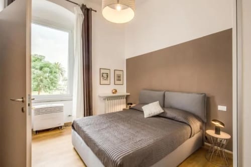 Camera da letto moderna con letto imbottito grigio, parete accent beige, finestra con luce naturale e arredi minimalisti