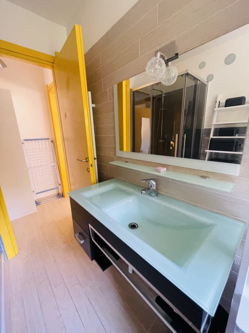 Modern bathroom with glass double sink, mirror, shower enclosure, and yellow door frame at Varenna by Foot hotel apartment