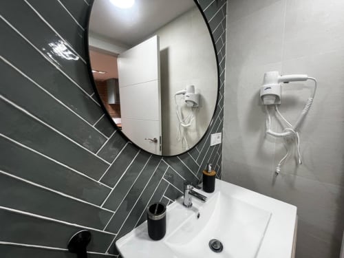 Baño moderno en ML Apartments Valencia con espejo curvo, lavamanos blanco y paredes de azulejos oscuros con dos apliques para secador