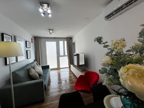 Sala de estar moderna en Valencia con butaca verde azulada, silla roja, pisos de madera, flores frescas y puertas de cristal