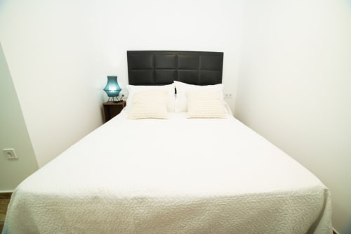 Dormitorio moderno con ropa de cama blanca, cabecero tapizado negro, lámpara turquesa y decoración minimalista en apartamento de Valencia