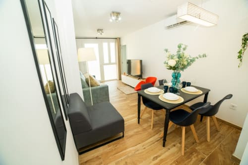 Comedor moderno de apartamento con mesa negra, suelos de madera y zona de estar junto a Playa Malvarrosa en Valencia.