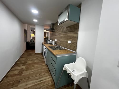 Cocina moderna con gabinetes color verde azulado, encimera de madera, fregadero, estufa, lavadora y silla alta blanca en ML Apartments