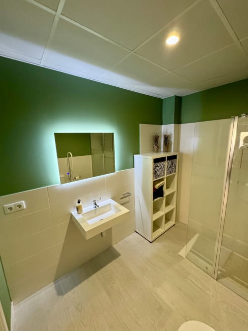 Baño moderno en ML Apartments Valencia con paredes verdes, espejo iluminado, azulejos blancos, ducha y estantes de almacenamiento