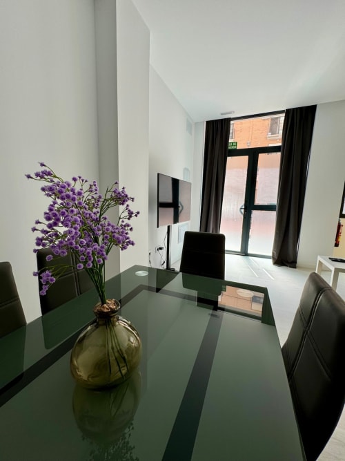 Zona de comedor moderno en ML Apartments Valencia con flores púrpuras, mesa verde y zona de trabajo junto a ventanas con cortinas negras