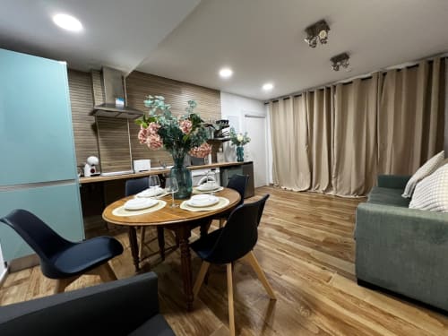 Comedor moderno con mesa ovalada de madera, sillas negras, refrigerador turquesa, flores frescas y cortinas beige