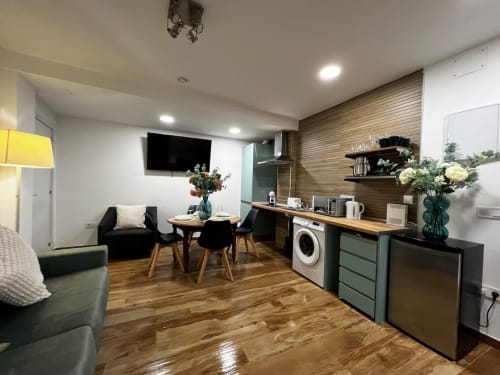 Sala de estar moderna en apartamento con cocina americana, sillas negras, pared de acentos de madera y TV en ML Apartments Valencia