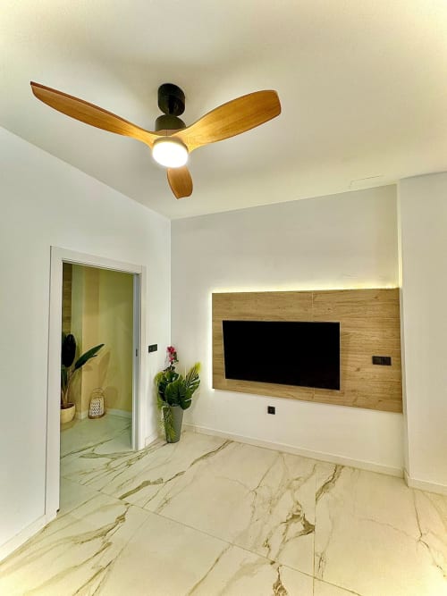 Sala de estar moderna con ventilador de techo de madera, piso de mármol, televisor montado en pared y plantas en maceta cerca de la puerta de entrada