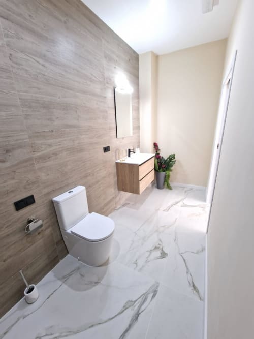 Baño moderno en apartamento de Valencia con piso de mármol, azulejos efecto madera, inodoro blanco, lavamanos y flores en maceta