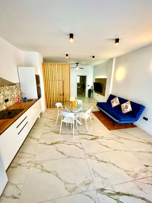Moderno apartamento diáfano en Valencia con suelo de mármol, mesa de comedor blanca, sofá azul, cocina equipada y techo