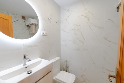 Modern bathroom in SUNSET BAY 509 at Home Stay Gran Canaria with marble walls, white sink, toilet, illuminated mirror, and