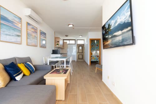 Modern open-plan living area in Sunset Bay apartment, Gran Canaria, with gray sofa, dining table, kitchen, TV, and sea view