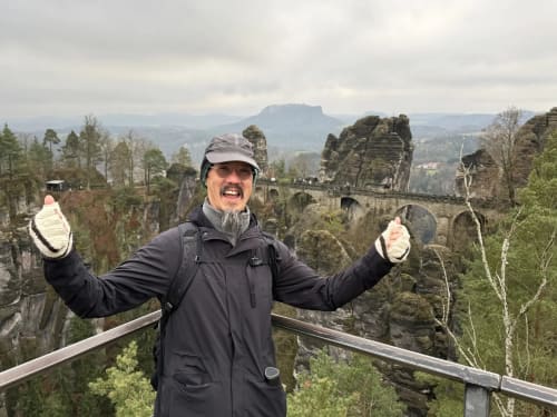 Man celebrating at Bastei Bridge viewpoint, Saxony Germany with dramatic sandstone cliffs and forest landscape behind him