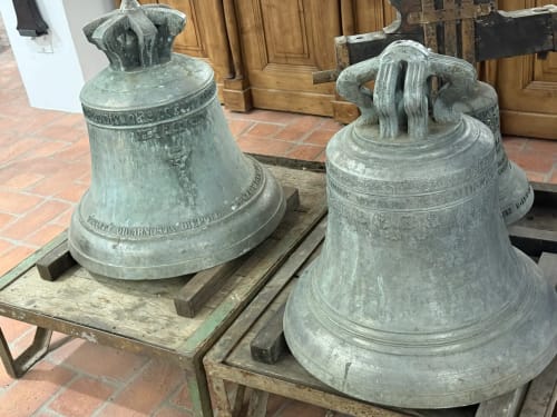 Two large bronze bells with ornate details on wooden stands in historic Ribeirinha courtyard setting.