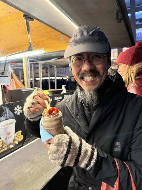 Man with beard smiling at Golden Mermaid Villa food stall in Ribeirinha, Portugal, holding wrapped sandwich