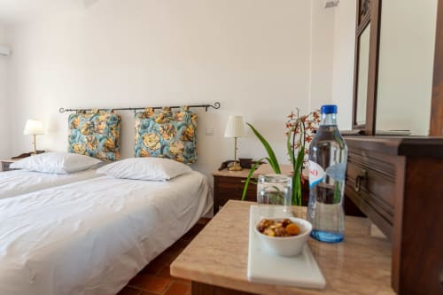 Quarto luminoso em Cerro da Campaniça com roupa de cama branca, almofadas estampadas florais, móveis de madeira e vista para o jardim