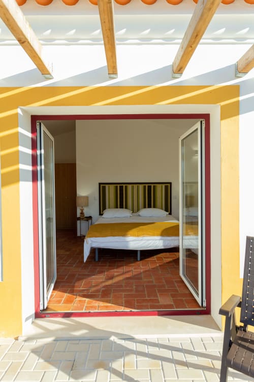 Quarto em Cerro da Campaniça com cama queen, vista para o jardim através de portas de correr, pisos em terracota e sombra de pérgula