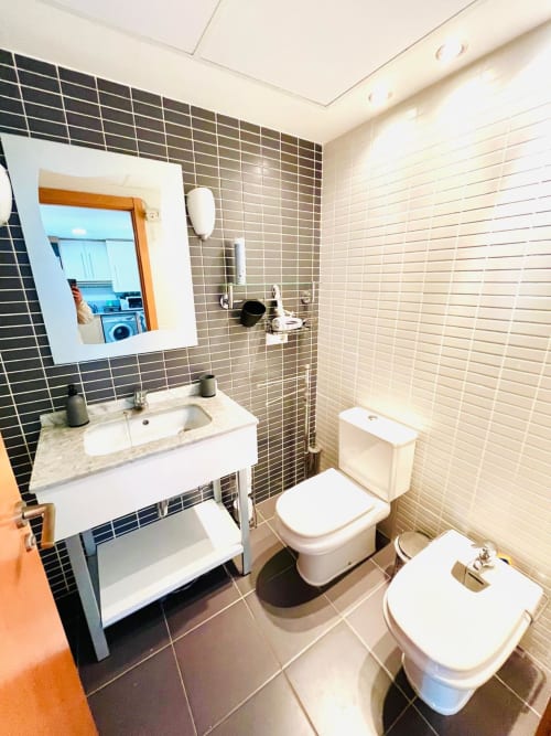 Salle de bain moderne aux murs en carrelage foncé, vasque blanche, toilettes et miroir dans l'appartement Sant Jordi
