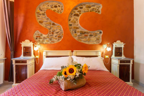 Camera da letto arancione vibrante a Casa Scola Loano con letto, composizione di girasoli e lettere SC in pietra sulla parete