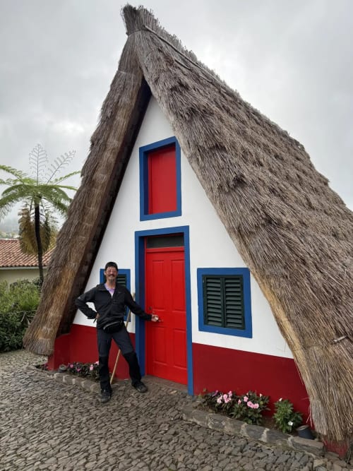 Homem posando na característica cabana em forma de A da Golden Mermaid Villa, com teto de colmo, acabamento azul e porta vermelha em Ribeirinha,