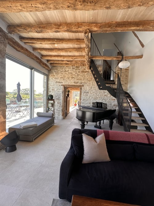 Spacieux séjour de villa avec poutres en bois apparentes, grand piano, murs en pierre, canapé noir et escalier au Gîte l'Harmonie