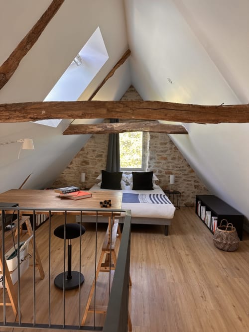 Chambre rustique en sous-pente avec poutres apparentes, mur en pierre, lit aux coussins noirs, bureau et lucarne à la Gîte