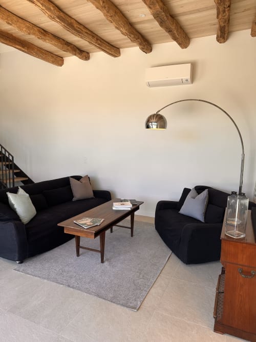 Salon moderne du gîte l'Harmonie du Causse avec canapés noirs, poutres en bois apparent, lampadaire arc et décor rustique