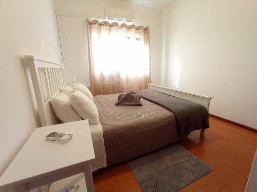 Quarto luminoso no Sweet Home Inn Portimão com cama de casal, móveis brancos e vista da cidade pela janela ensolarada