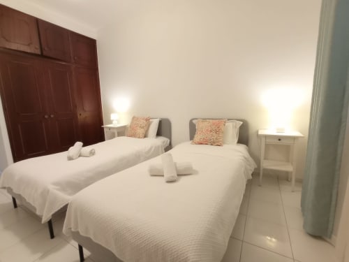 Quarto duplo no hotel Sweet Home em Portimão com camas brancas, almofadas coral, guarda-roupa escuro e mesas laterais com luminárias