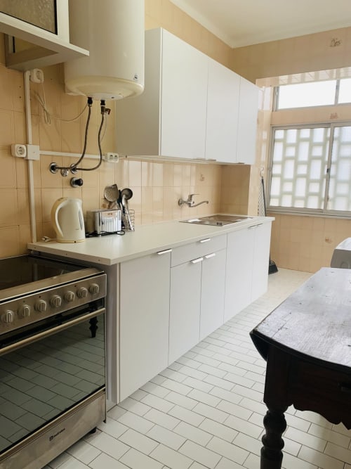 Cozinha do quarto do hotel Sweet Home em Portimão: armários brancos, fogão de aço inoxidável, azulejos bege, luz natural da janela
