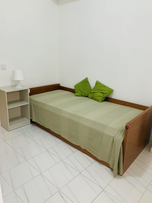 Quarto luminoso no Sweet Home Portimão com cama de madeira, almofadas verdes, criado-mudo branco e luminária em piso de azulejos brancos