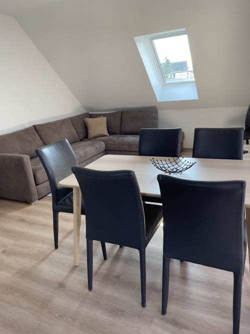 Salle à manger aménagée dans les combles avec chaises noires, table blanche, canapé brun, puits de lumière et parquet chez La clé des vignes