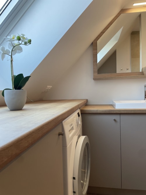 Buanderie mansardée moderne dans l'appartement La clé des vignes à Ribeauvillé, avec lave-linge blanc, comptoir en bois et finitions blanches