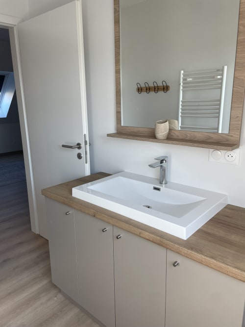 Salle de bain moderne avec lavabo blanc rectangulaire, plan de travail en bois, sèche-serviettes chauffant et crochets décoratifs à La clé des vignes
