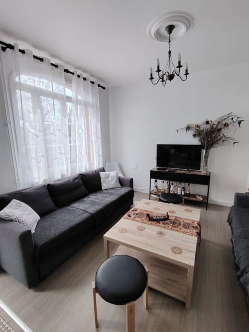 Séjour contemporain du gîte Skol In Normandy à Rouen avec canapé noir, table basse en bois, téléviseur écran plat et voilages
