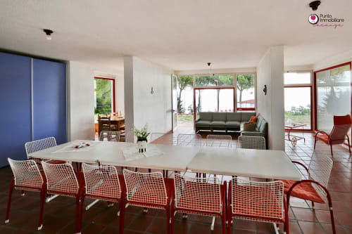 Modern villa dining area with sea views, featuring long white tables, red patterned chairs, and open-plan living space in