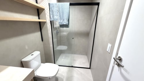 Banheiro moderno com chuveiro em vidro, vaso sanitário, pia e prateleiras de madeira no quarto 211 da pousada em Brasília