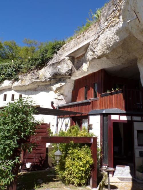Les Troglos de Beaulieu troglodyte room entrance with red wooden doors, stone cliff facade, and garden seating area in