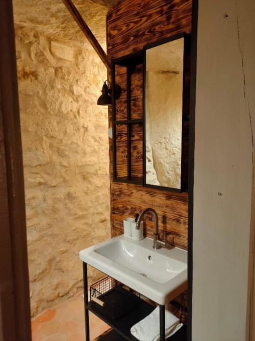 Salle de bains rustique avec lavabo blanc, miroir encadré de bois et murs en pierre aux Troglos de Beaulieu