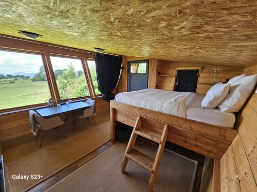 Chambre de cabane en bois à Logis Las Crozas avec grandes fenêtres donnant sur la campagne verdoyante, lit en bois avec literie blanche