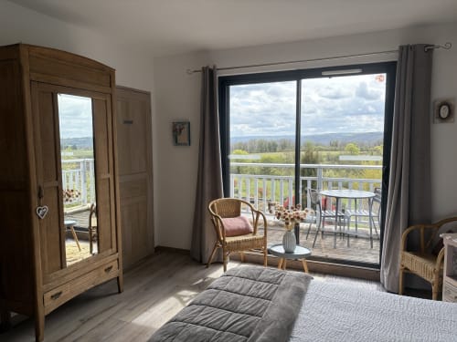 Chambre lumineuse aux Étoiles de Calès avec penderie en bois, fauteuil en rotin et portes vitrées ouvertes sur la campagne