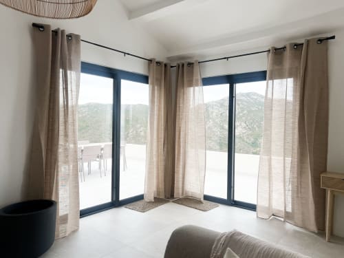 Chambre d'hôtel moderne avec baies vitrées du sol au plafond, rideaux beiges et vue sur les montagnes à Saint-Florent, Corse.