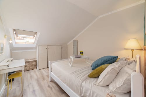 Quarto luminoso no sótão da pousada em Lisboa com camas de casal, almofadas azuis e amarelas, claraboias, guarda-roupas brancos e área de trabalho.
